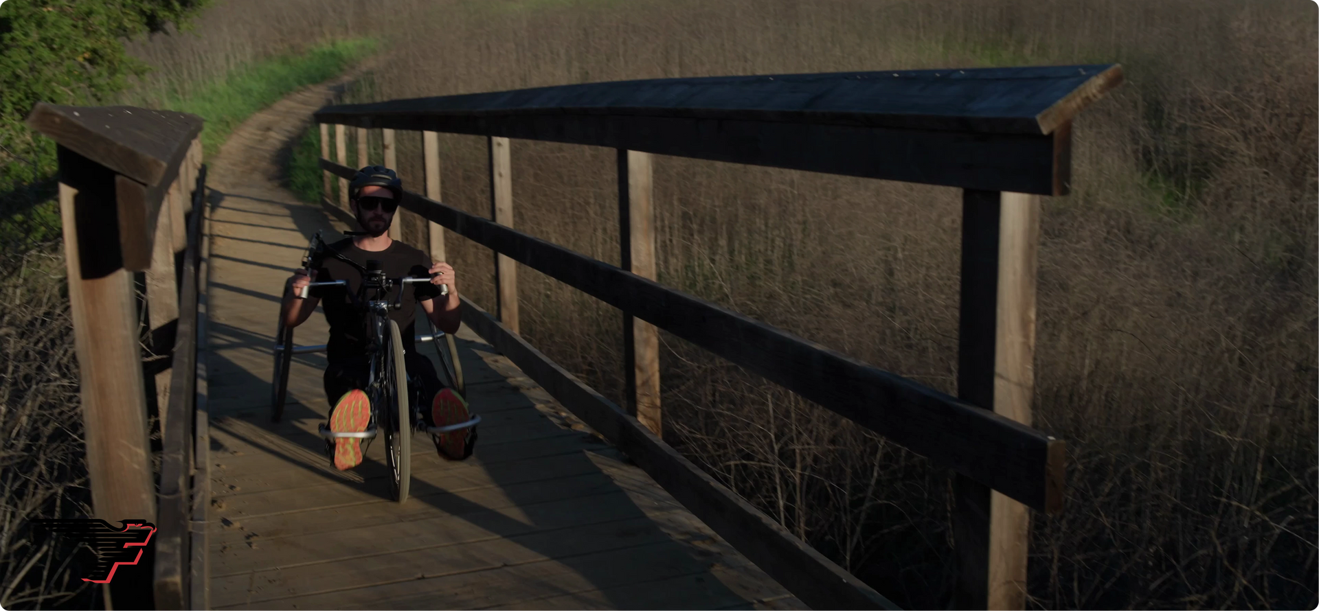 A man in a helmet and sunglasses rides a black handcycle across a wooden bridge on a nature trail.