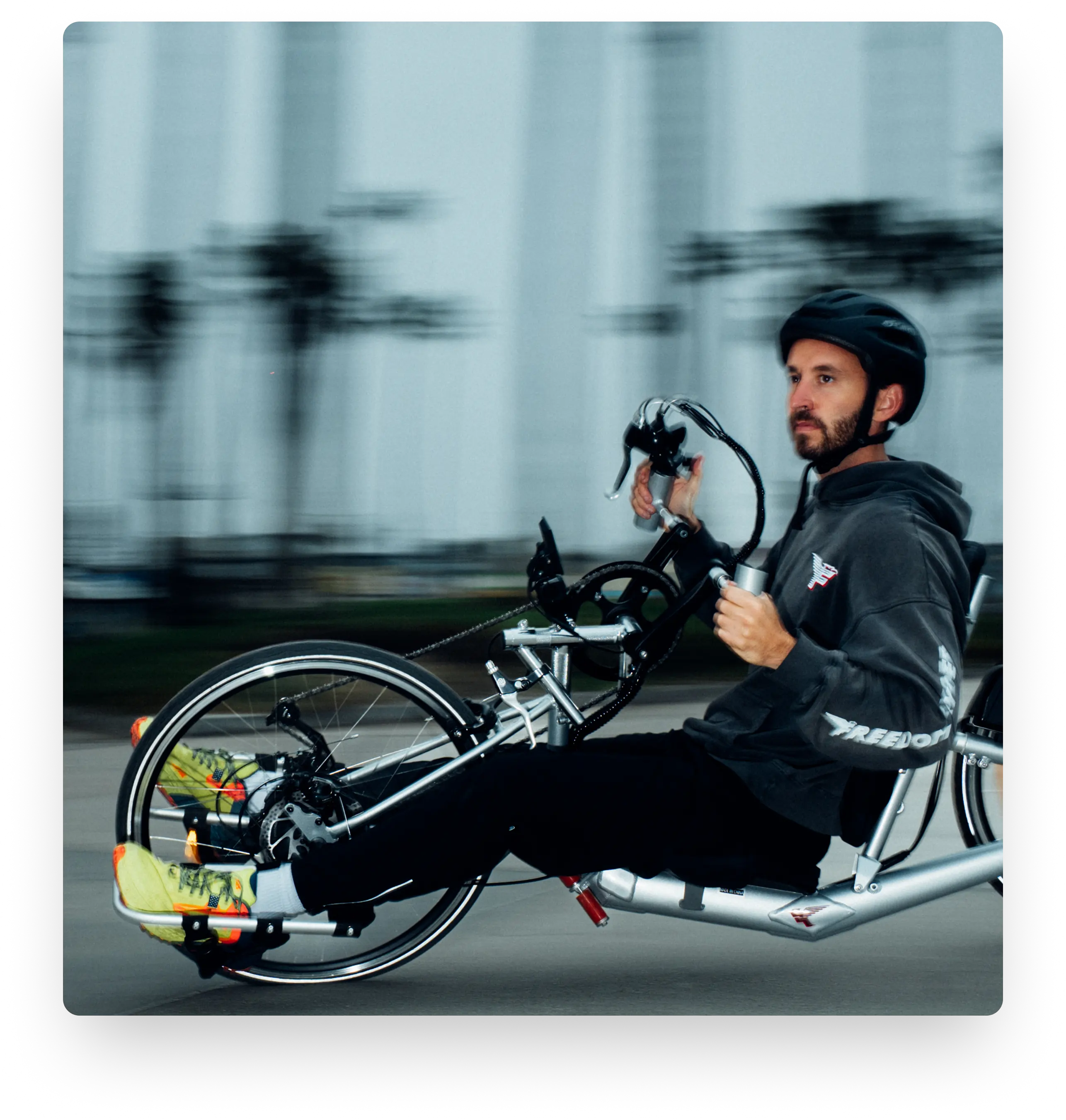 A man in a black helmet and grey hoodie riding a silver Freedom Ryder handcycle at high speed.