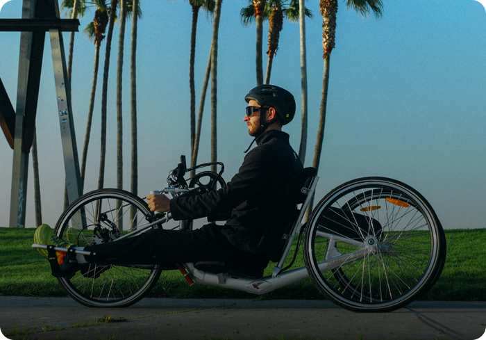 Person sitting in a recumbent handcycle on a sunny path lined with palm trees during initial consultation