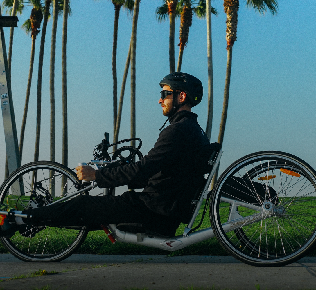 Person riding an E1 handcycle outdoors with palm trees in the background