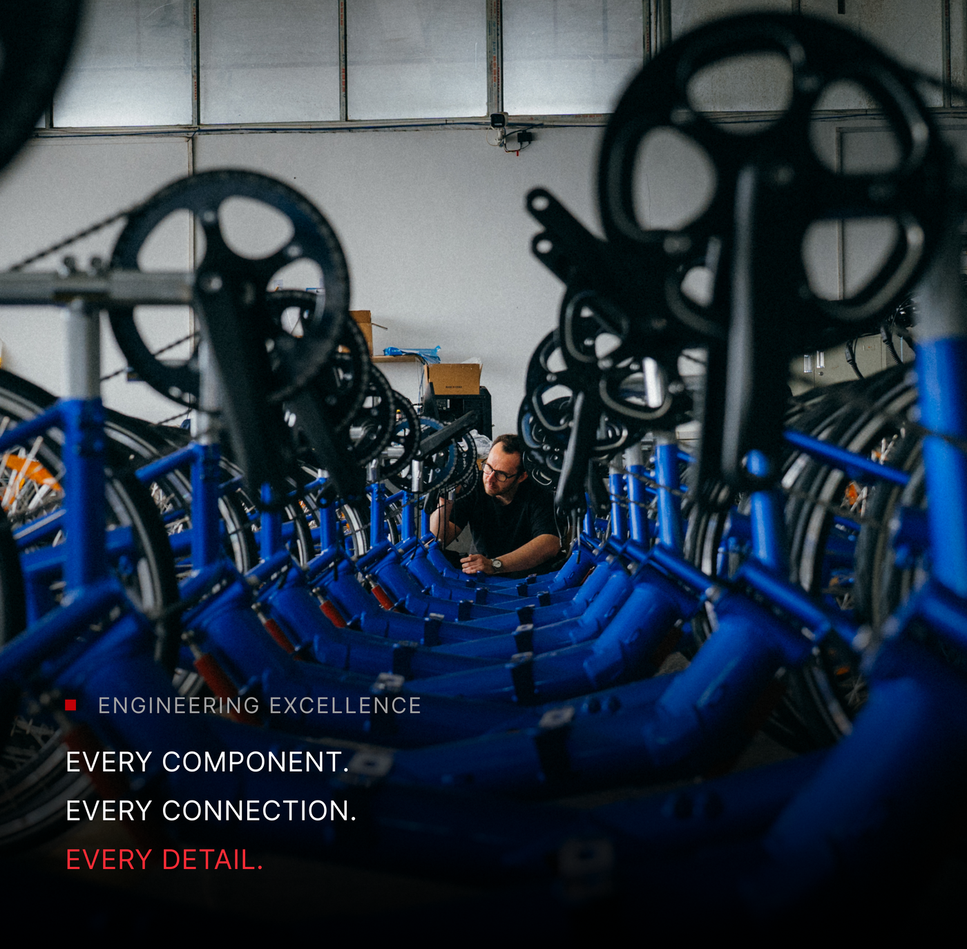 A technician works on a long row of blue bike frames in a warehouse, with text overlay reading "Engineering Excellence. Every component. Every connection. Every detail.