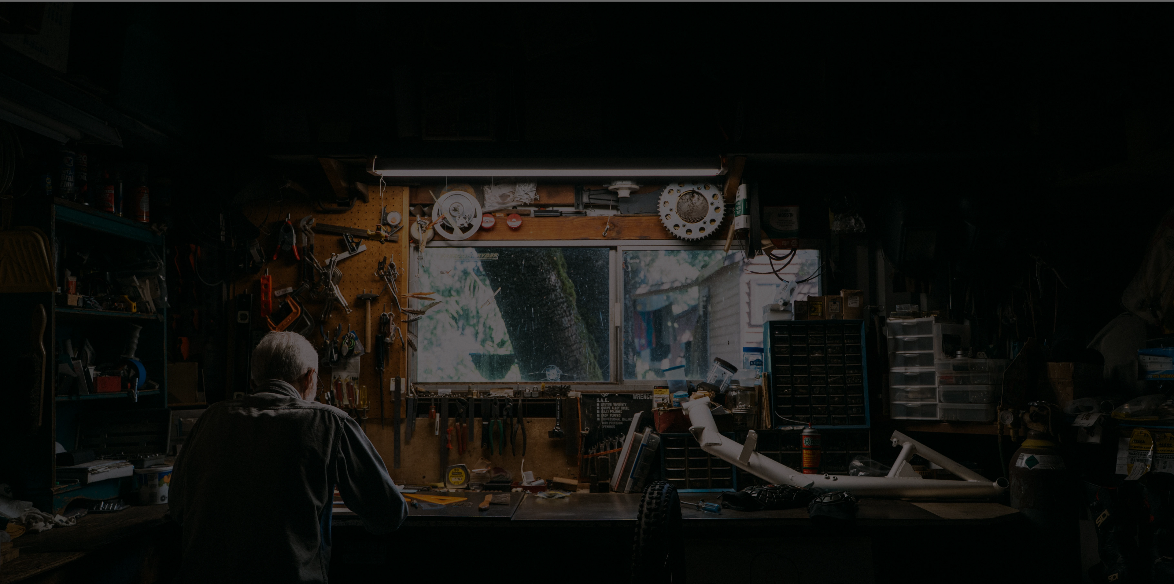 A man works at a cluttered workbench in a dimly lit garage, with a handcycle frame visible to the side.
