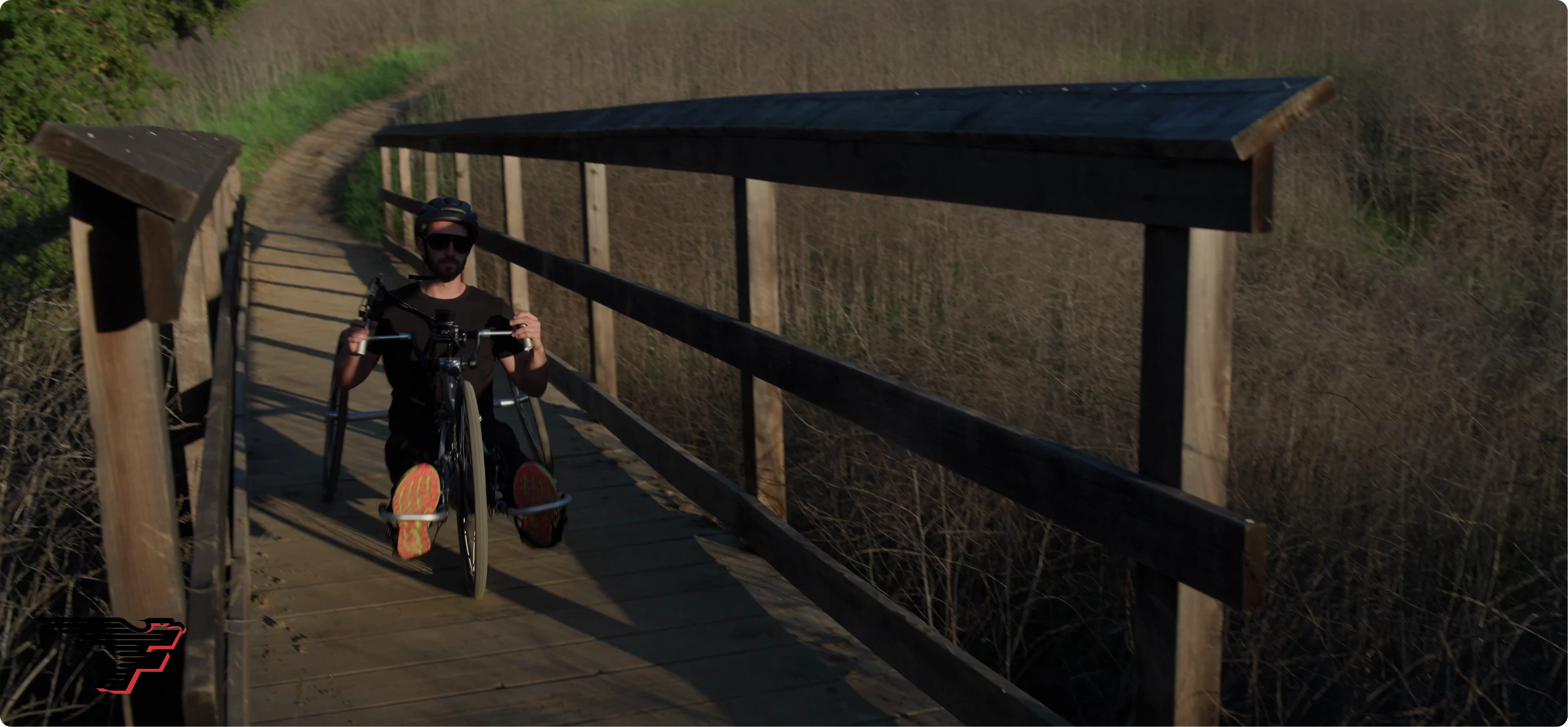 A man in a helmet and sunglasses rides a black handcycle across a wooden bridge on a nature trail.
