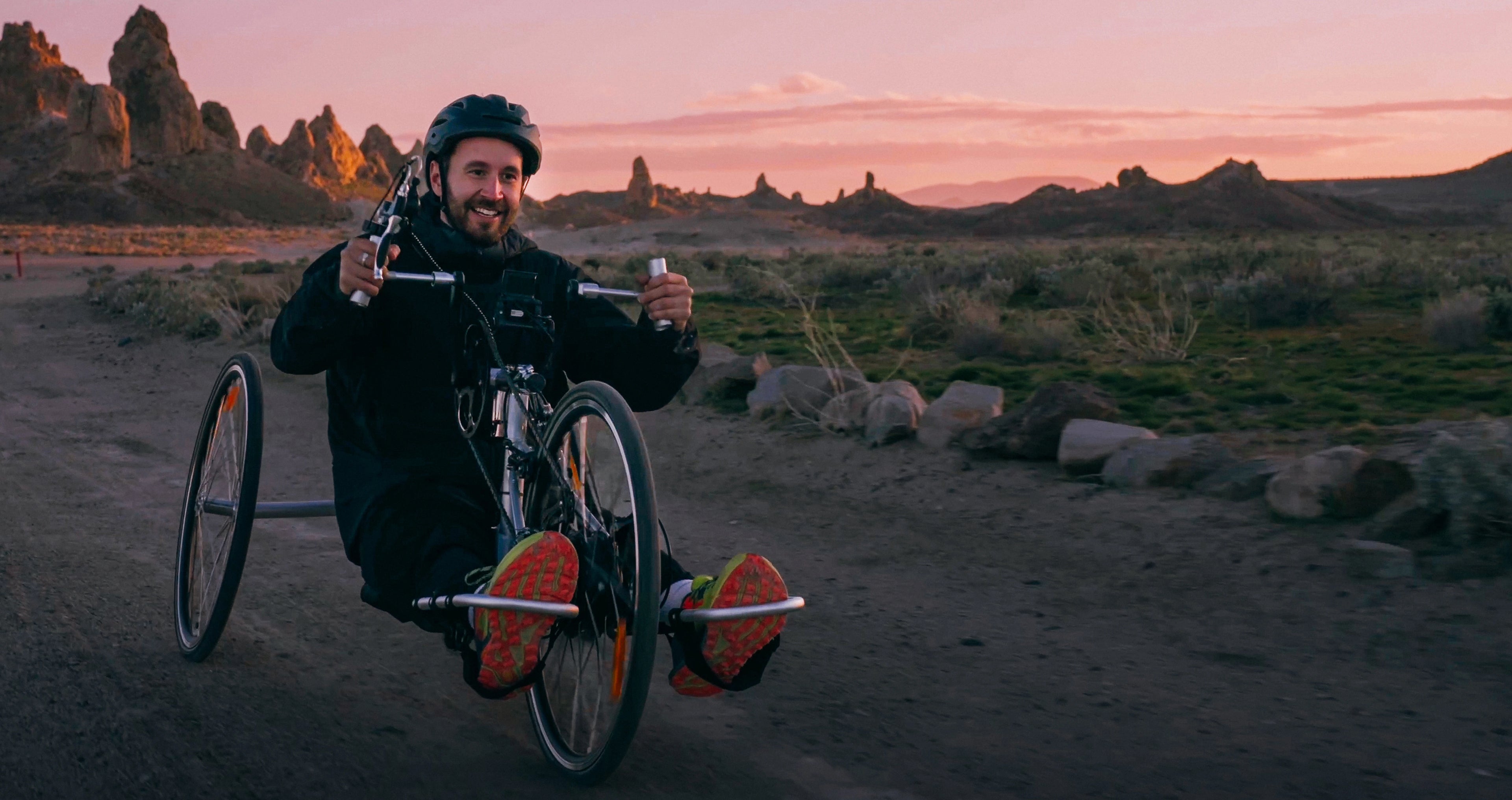 A smiling man in a helmet rides a silver handcycle down a desert path during a pink and orange sunset.