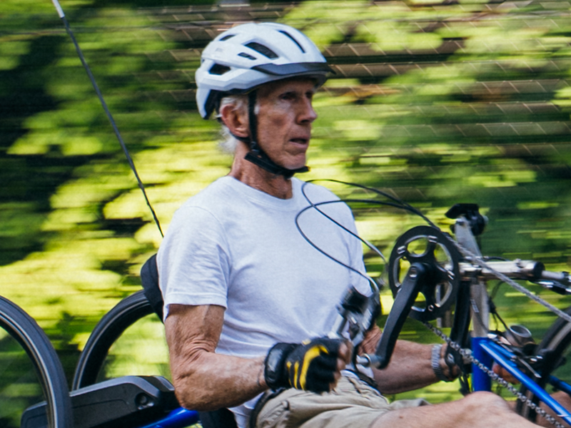 Elderly man riding a blue handcycle through a green outdoor trail wearing a white helmet