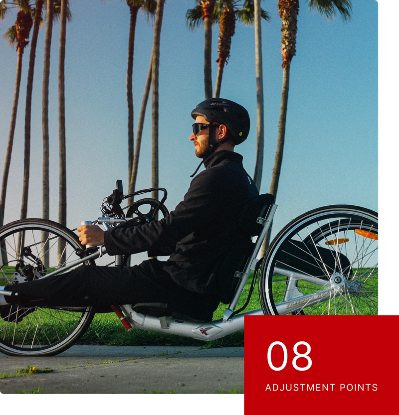 A man in a helmet and sunglasses rides a silver handcycle past a row of palm trees.